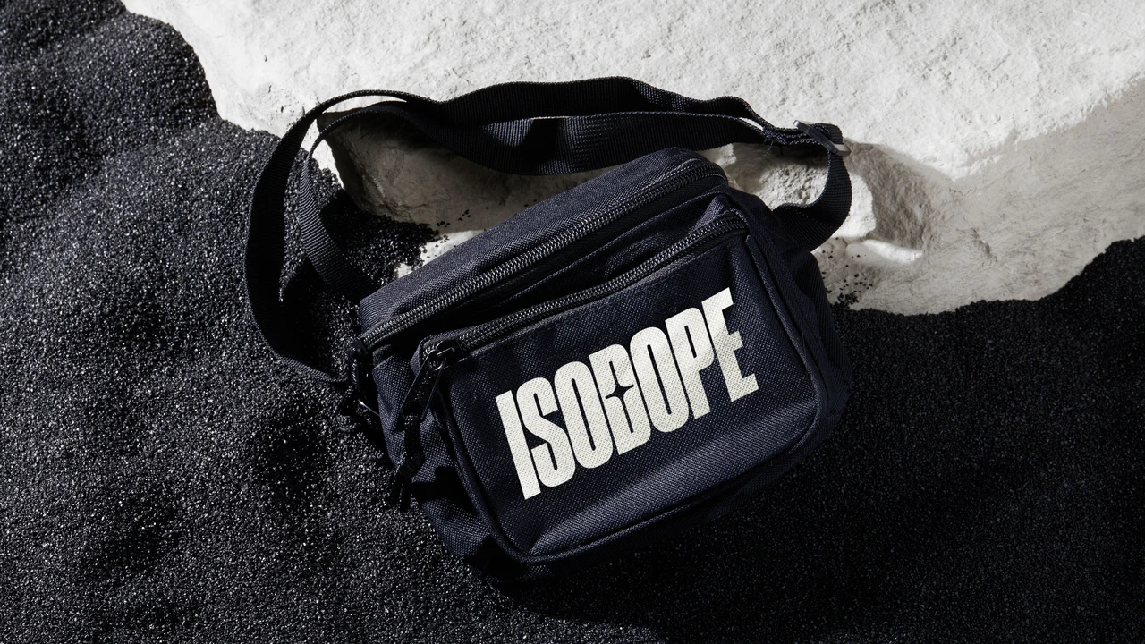 Bolsa transversal preta com logotipo Isodope branco é apresentada sobre terreno texturizado em preto e branco, com iluminação lateral forte.