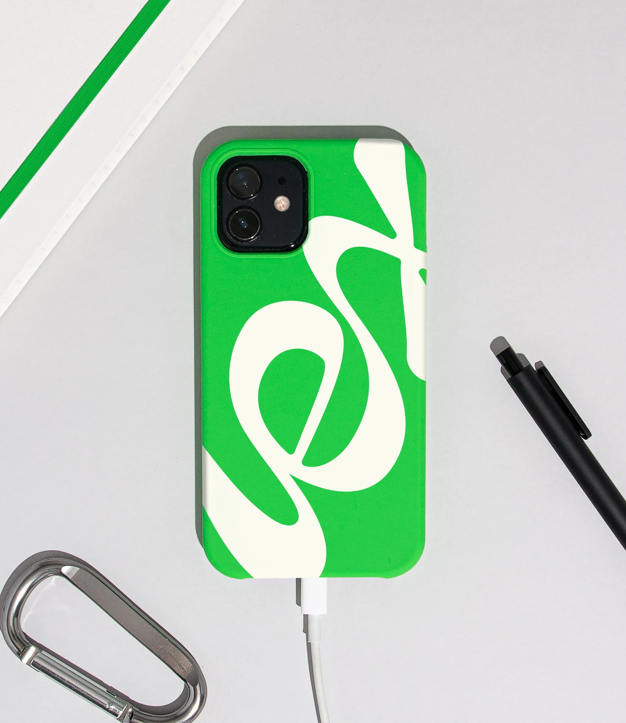 A green phone case carries an oversized cream Lex wordmark, with a white charging cable, pen and carabiner arranged nearby.