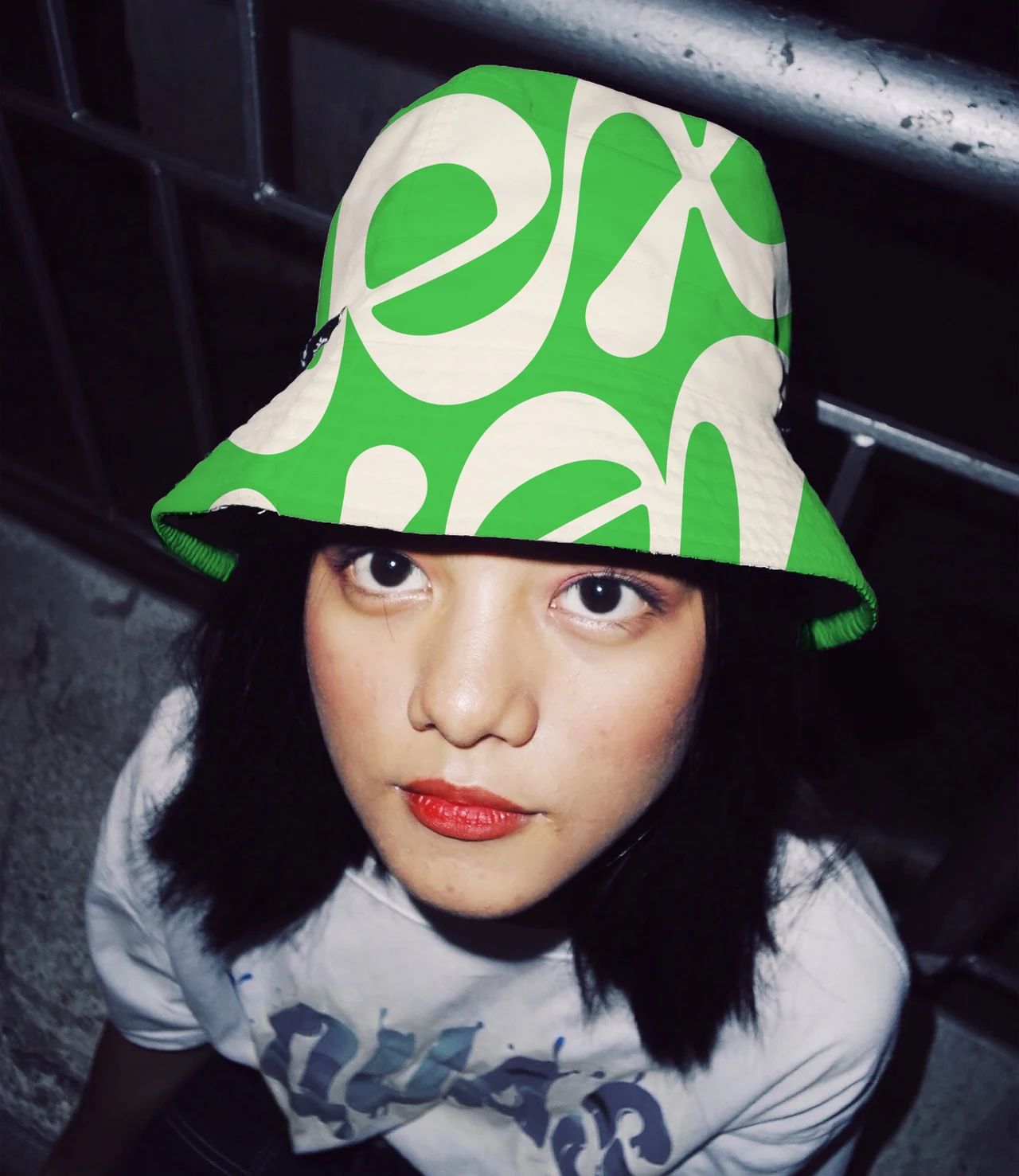 A high-angle flash portrait shows a person wearing a green and cream bucket hat patterned with oversized Lex letterforms.