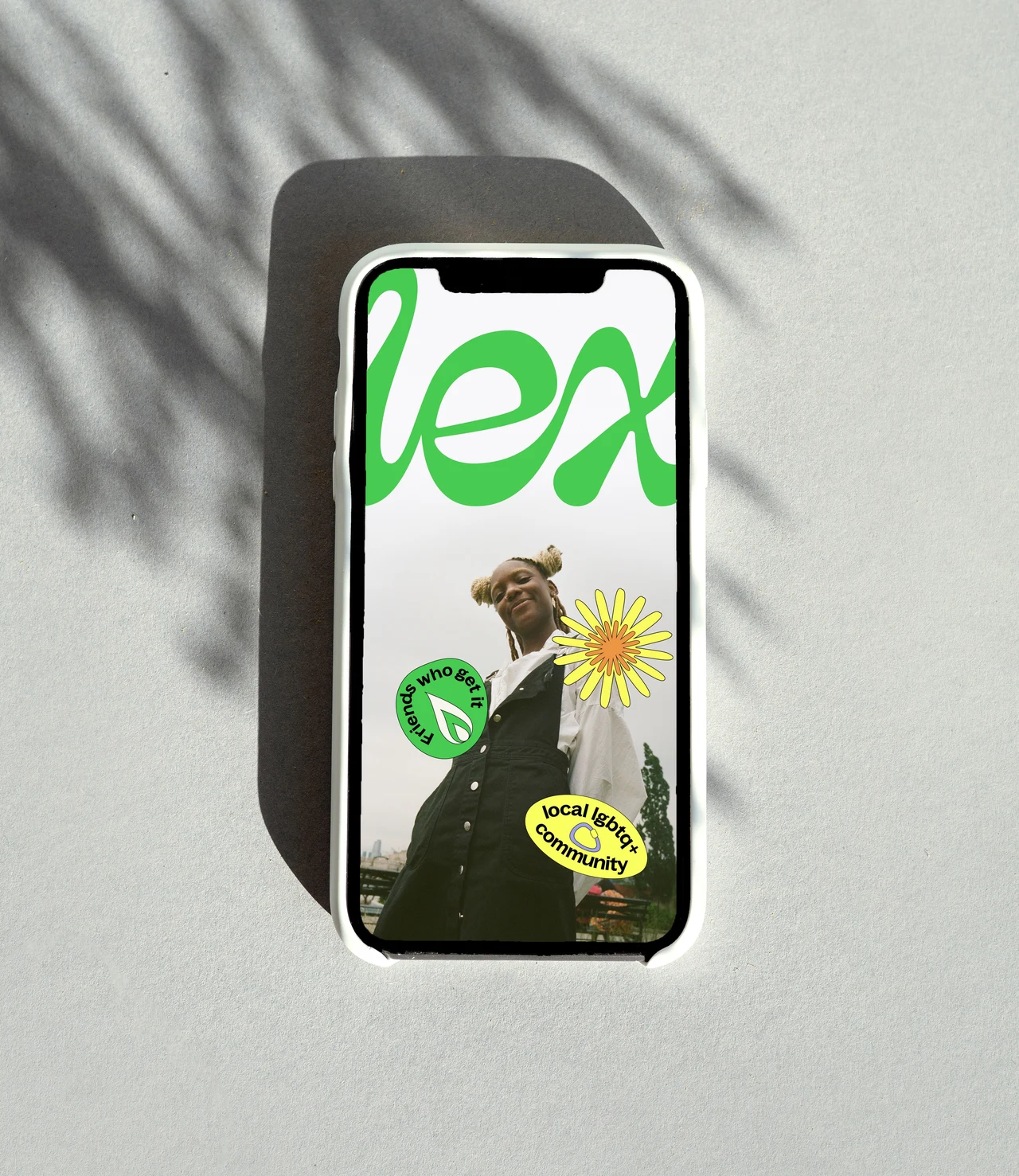A phone on a gray surface displays a vertical composition of a green Lex wordmark, portrait and yellow and green stickers under leaf shadows.