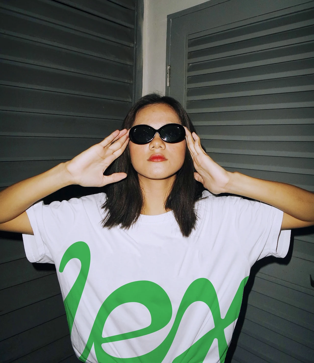 A person in sunglasses poses in front of a metal door wearing a white T-shirt with an oversized green Lex wordmark.