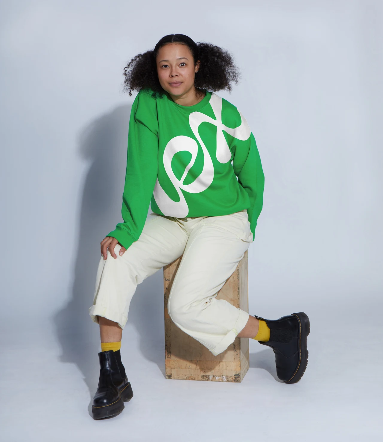 A full-length portrait shows a person seated on a wooden block wearing a green Lex sweatshirt, pale trousers and black boots.