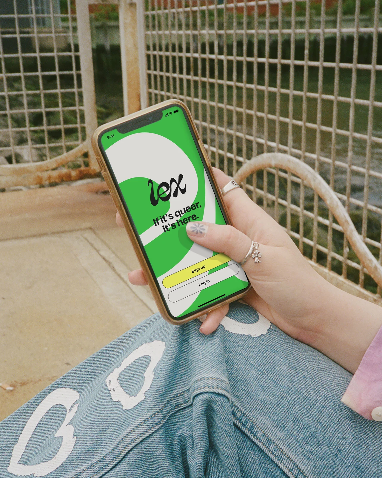 A hand holds a phone showing the green Lex welcome screen above heart-decorated jeans and a metal fence.