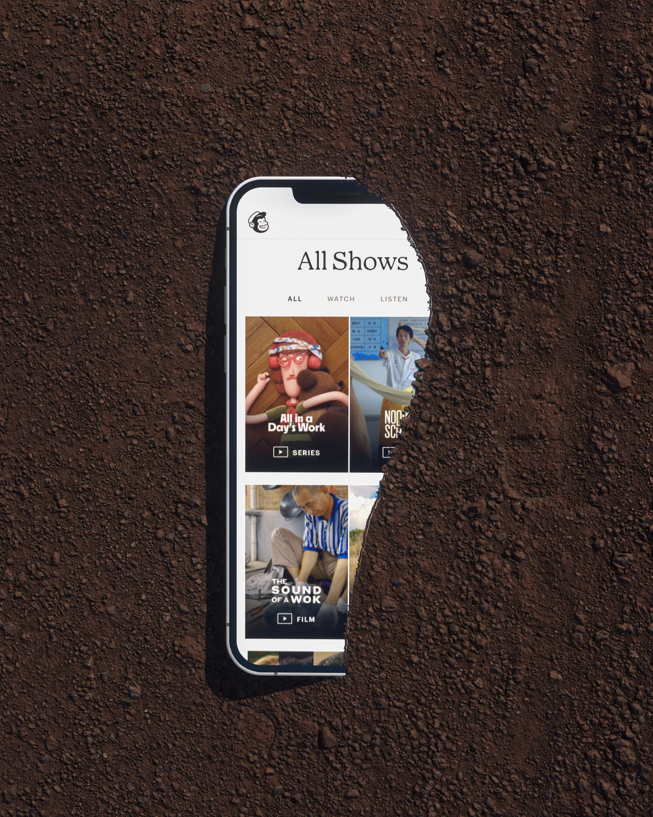 A phone mockup with Mailchimp’s program catalog sits inside an irregular opening in dark earth.