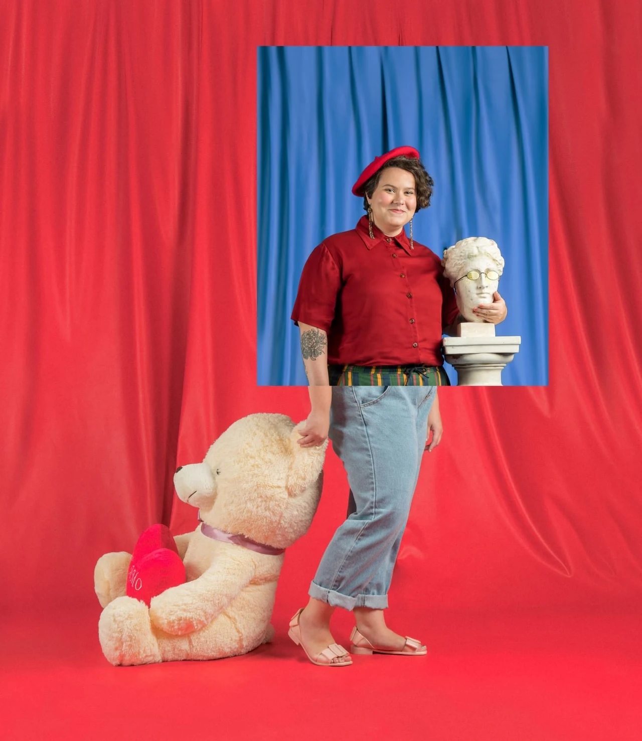 Pessoa de boina vermelha posa descalça com urso de pelúcia e busto clássico, diante de cortinas vermelha e azul.