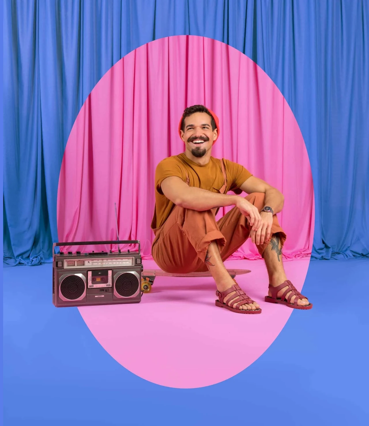 Pessoa sorridente sentada junto de um rádio portátil aparece em cenário azul e rosa, com cortinas e um grande recorte oval.