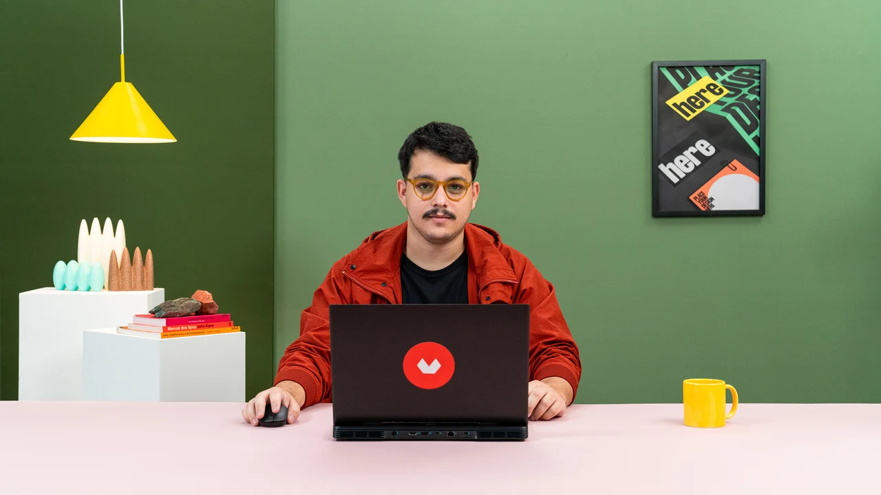A presenter in a red jacket sits behind a laptop in a green studio with a pink table, a yellow pendant lamp and colorful objects.