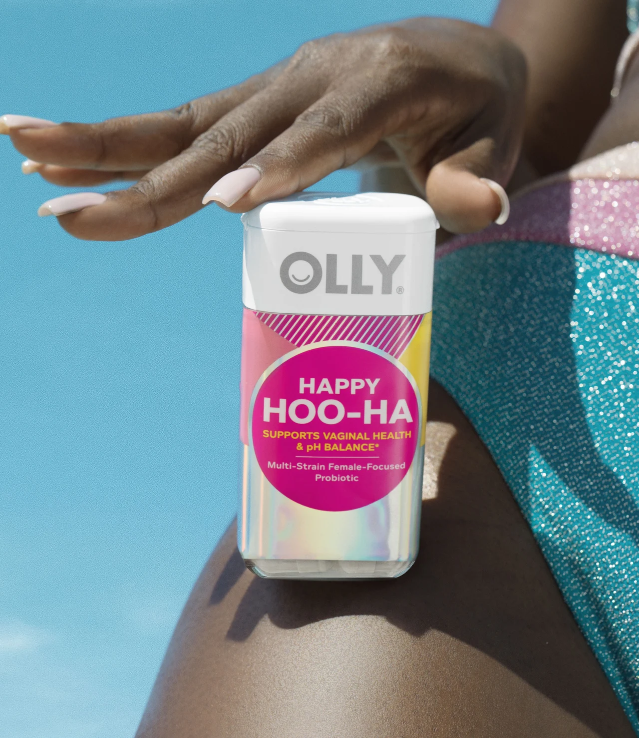 Close-up of a hand beside an OLLY Happy Hoo-Ha bottle resting on the thigh of a person in a glittery turquoise and pink swimsuit.