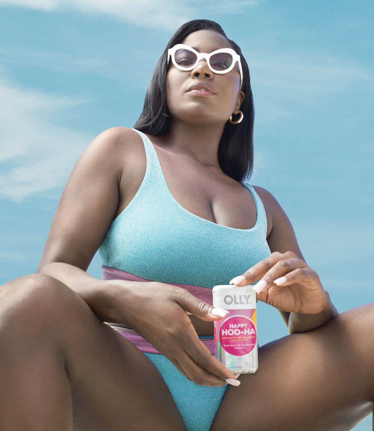Woman photographed from a low angle wears white sunglasses and a turquoise swimsuit while holding a Happy Hoo-Ha bottle against blue sky.