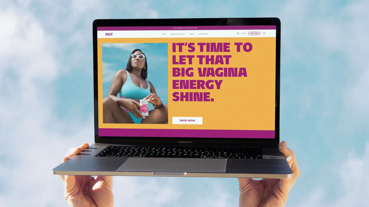 Laptop held by hands against a pale sky displays an OLLY page with navigation, a Shop Now button, swimsuit photography and oversized magenta text on yellow.