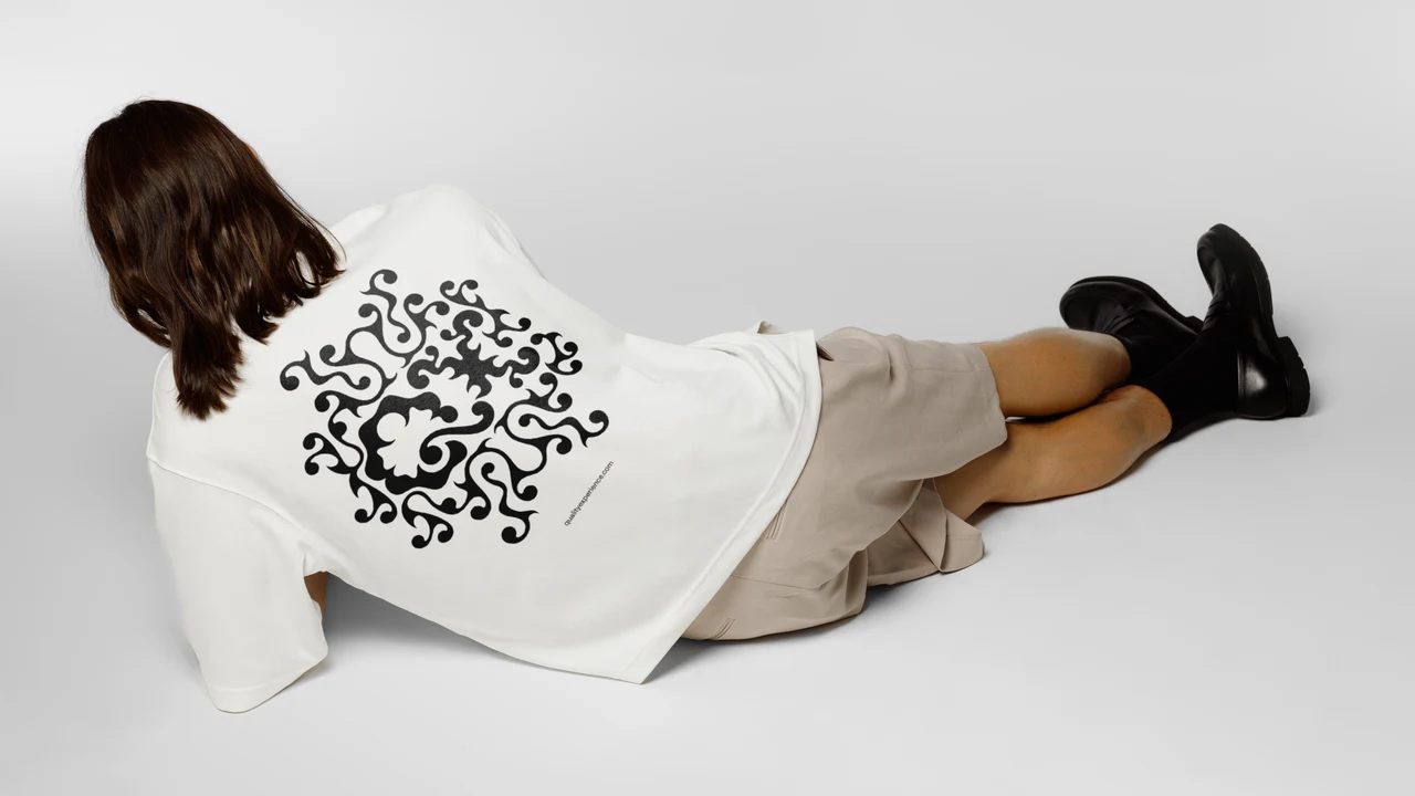 Person lying sideways in a studio wears an oversized white shirt with a circular black QX ornamental print, beige shorts and black boots.