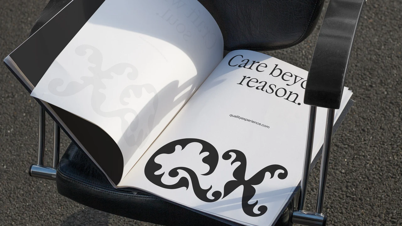 Open publication on a black chair shows a large QX monogram, serif “Care beyond reason” text and pale gray ornaments on white pages.