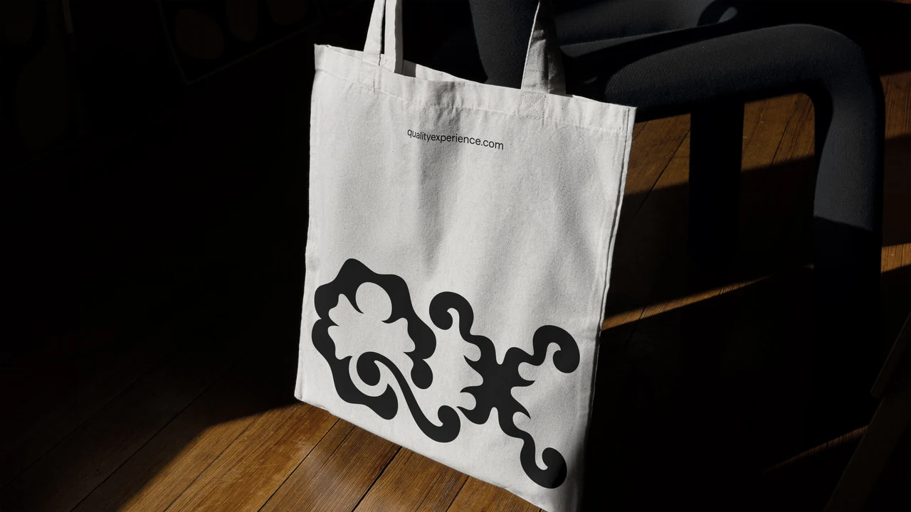 Pale cotton tote hanging from a wooden chair features a large black QX monogram at the bottom and a small address near the top.