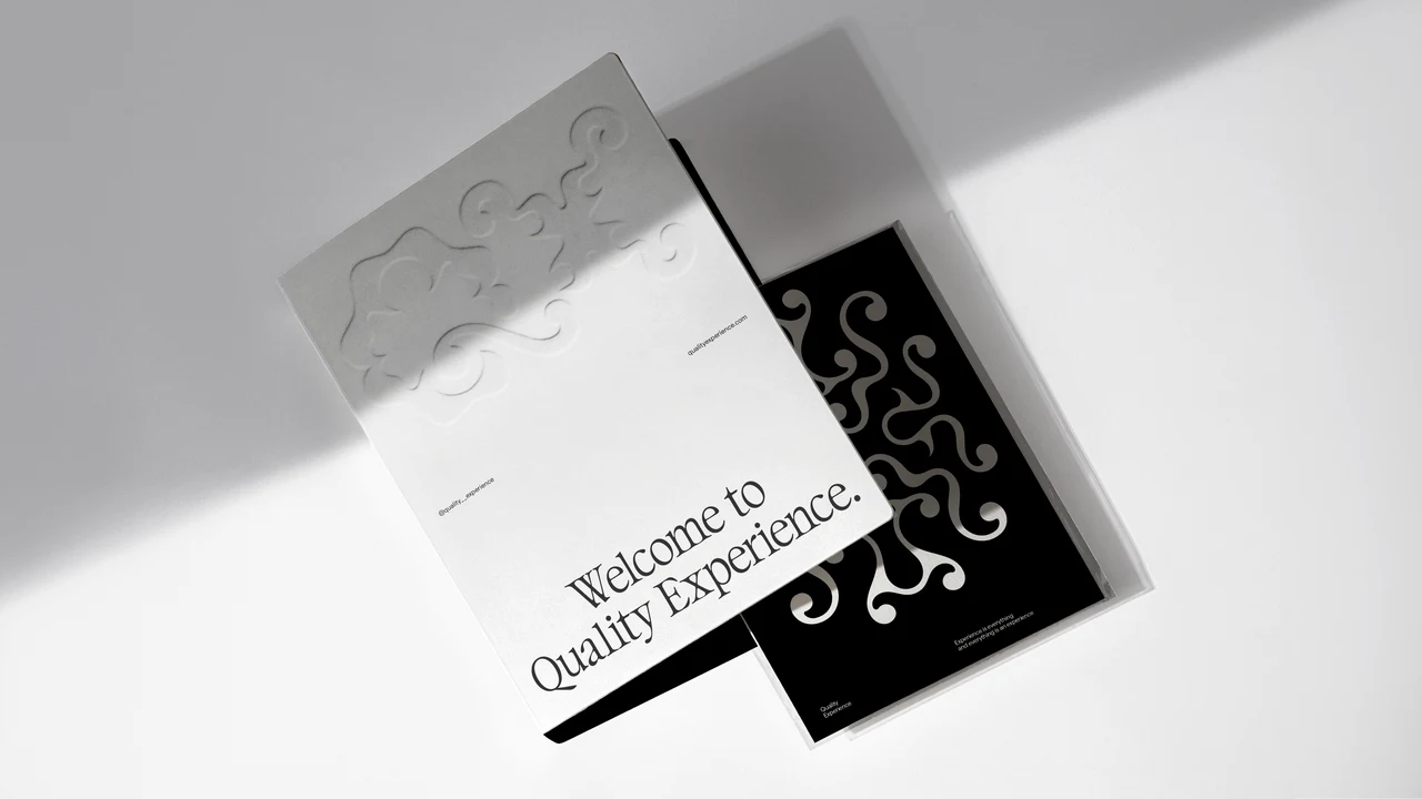 Two QX printed pieces overlap on a pale surface; the white piece has an embossed logo and “Welcome to Quality Experience”, while the black one shows an ornamental pattern.