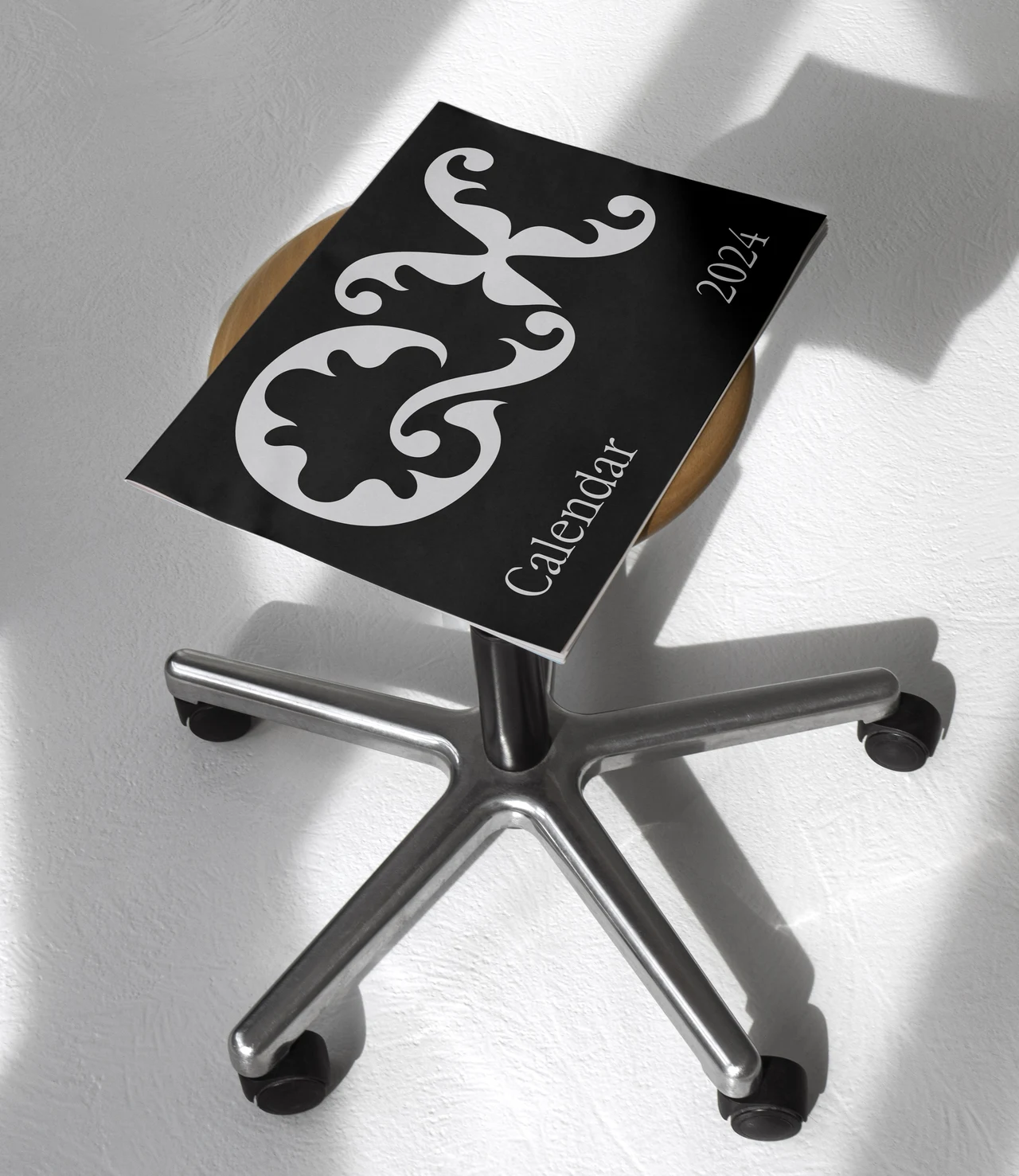 Black calendar with a large pale QX monogram and 2024 marking rests on a metal swivel chair seat in sunlight.