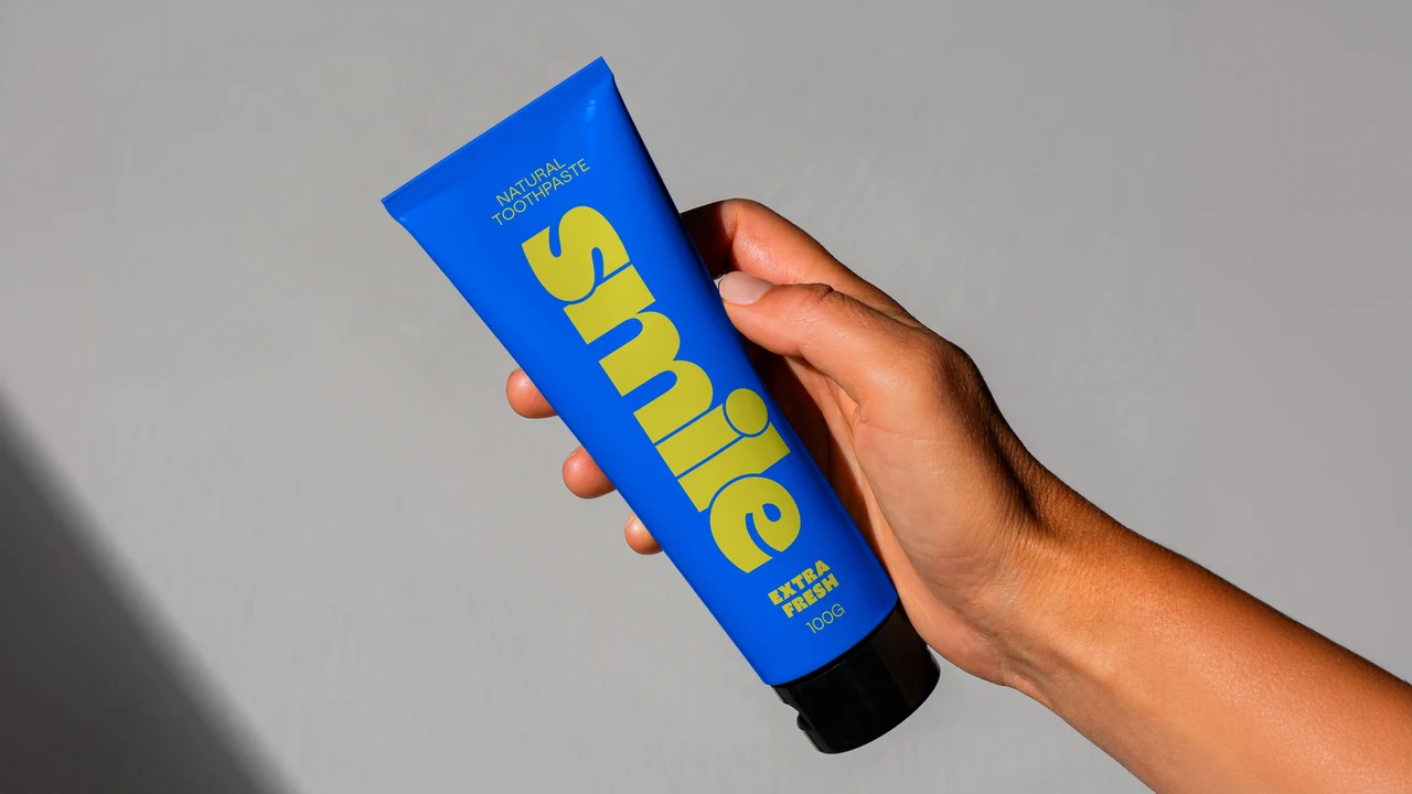 A hand holds a blue tube with a black cap and “smile” in rounded yellow lettering, photographed against gray with a diagonal shadow.