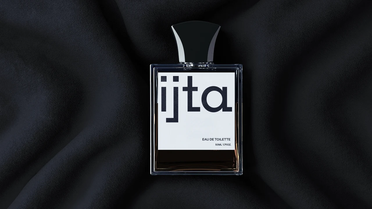 Rectangular perfume bottle with a sculptural black cap rests on dark fabric; its white label carries a name in geometric typography and “Eau de toilette” text.