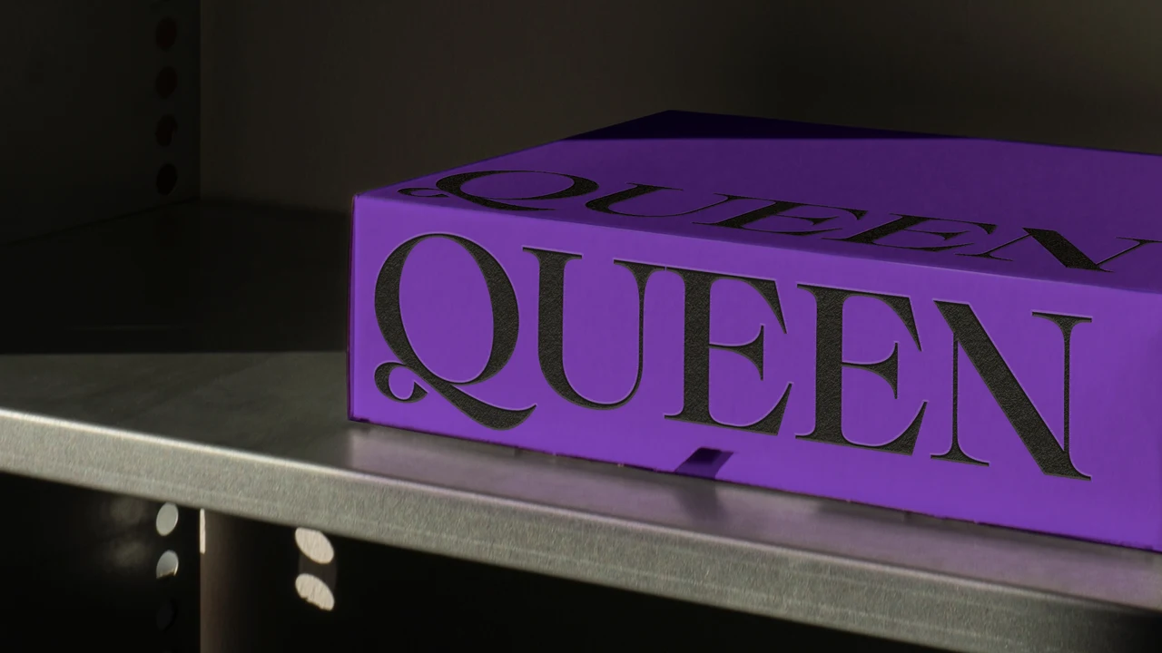 Purple box on a metal shelf displays “QUEEN” in very large black serif lettering, repeated on the lid.