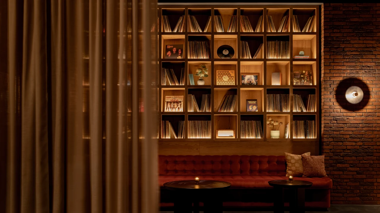 Lounge with warmly lit shelves of records and album covers, a red tufted banquette, small candlelit tables, a brick wall with a round sconce and a curtain in the foreground.