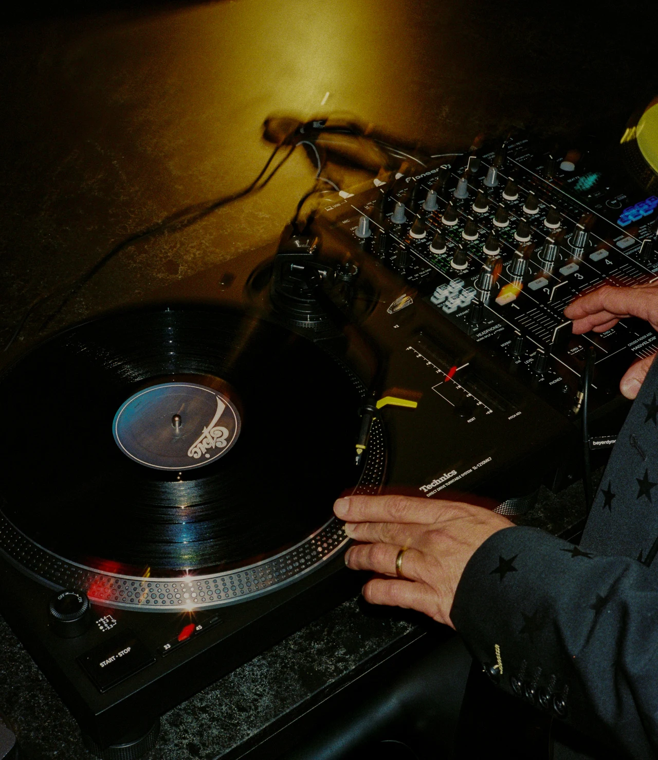 Flash photo of a DJ's hands on a Technics turntable and mixer, the record spinning under warm light.