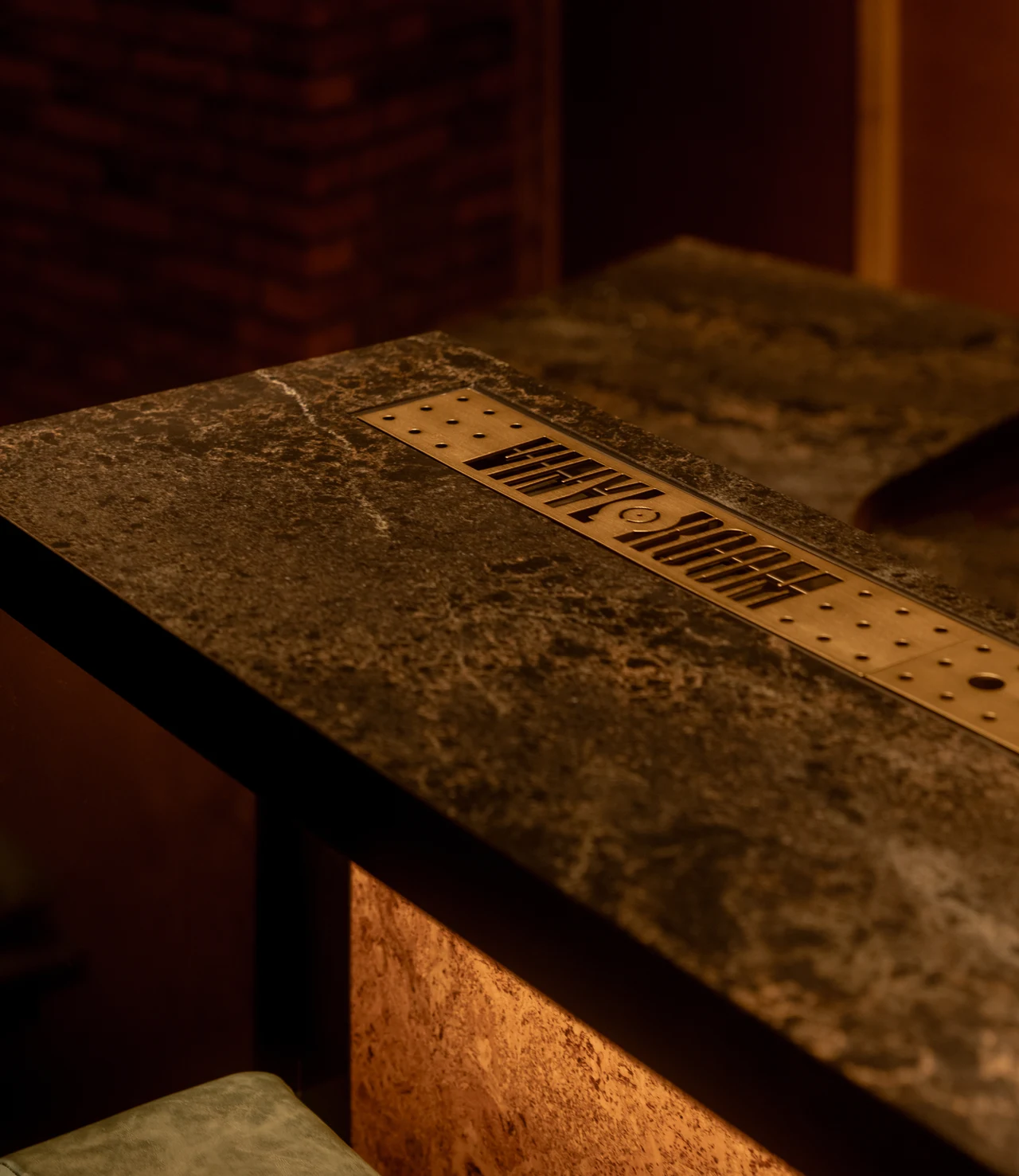 Perforated brass plate engraved with the VINYL ROOM wordmark, inset in a dark marble bar top under warm light.