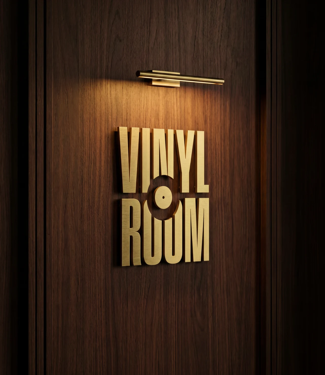 Brushed brass VINYL ROOM wordmark mounted on a dark wood wall under a brass picture light.