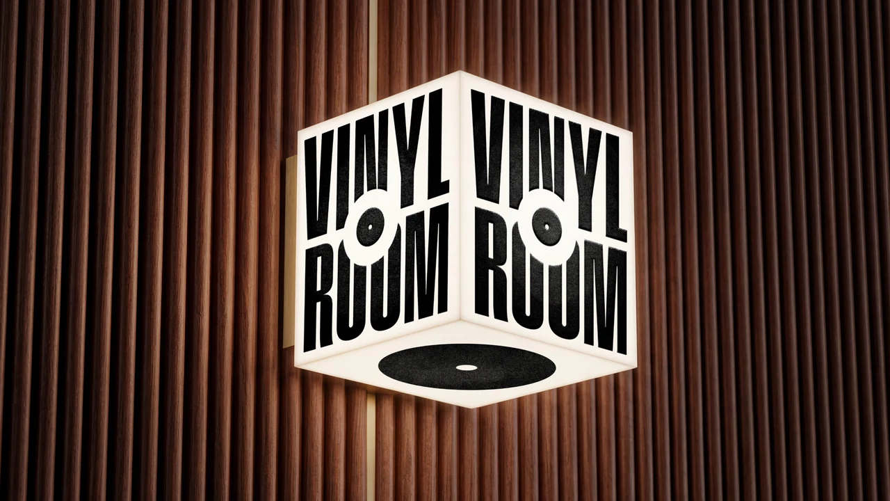 Illuminated cube sign with the black VINYL ROOM wordmark on its faces and a record on the base, mounted on a fluted wood wall.