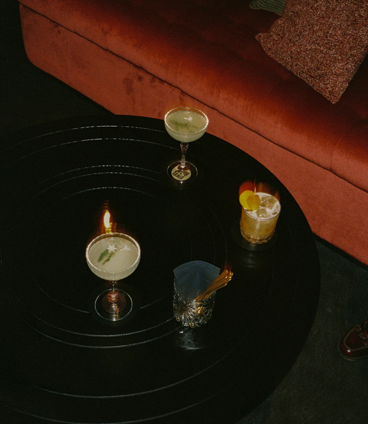 Flash photo from above of cocktails, a candle and a glass with gold straws on a round black record-like table beside a rust velvet sofa.