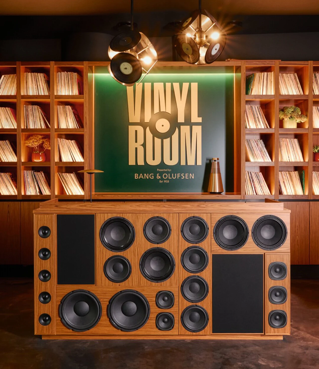 DJ booth with a wooden console full of speaker cones before a green panel with the gold VINYL ROOM wordmark presented by Bang & Olufsen, flanked by record shelves and record-shaped pendant lights.