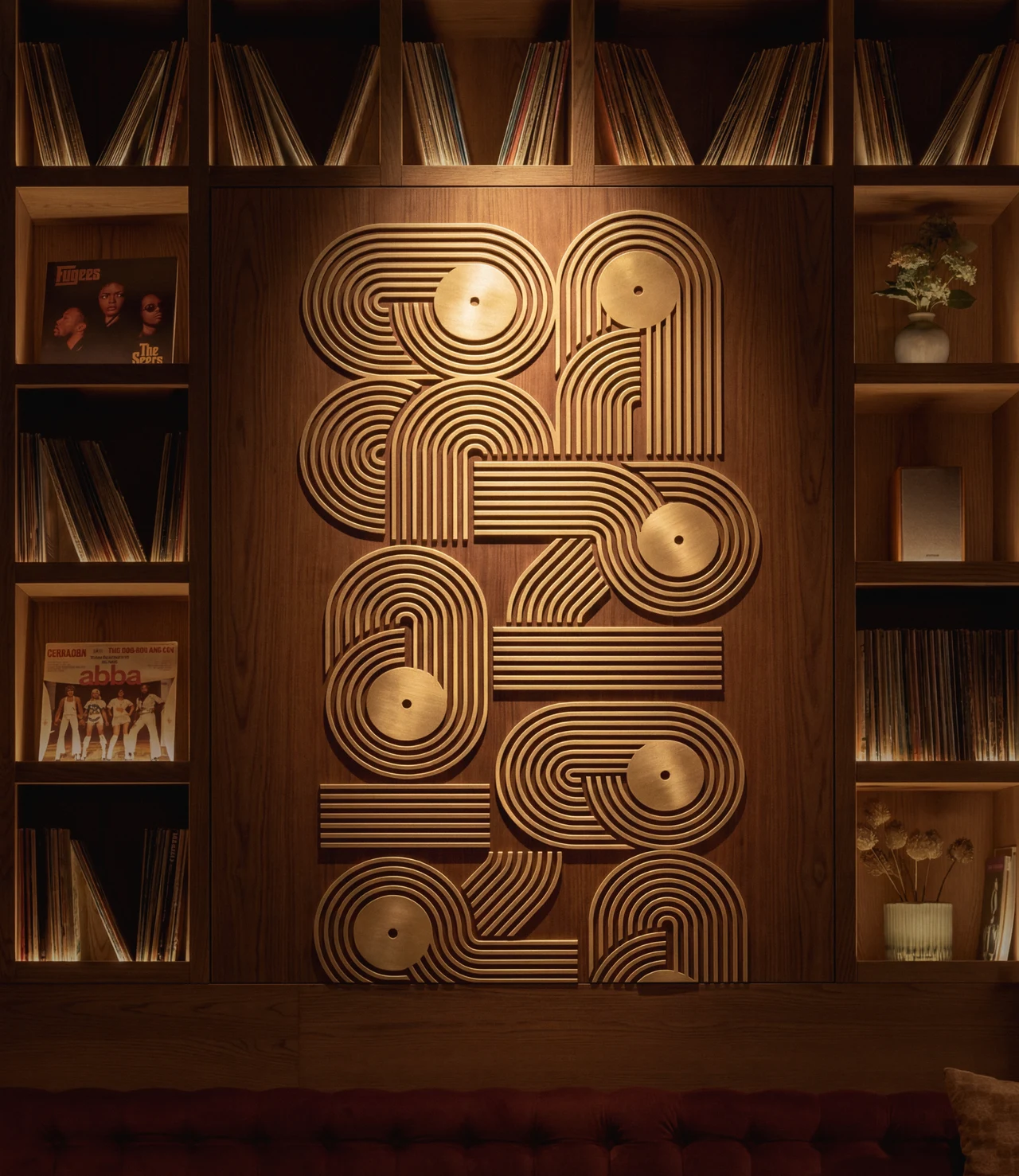 Brass wall installation of looping concentric lines and record discs, set between shelves of records and album covers in warm light.