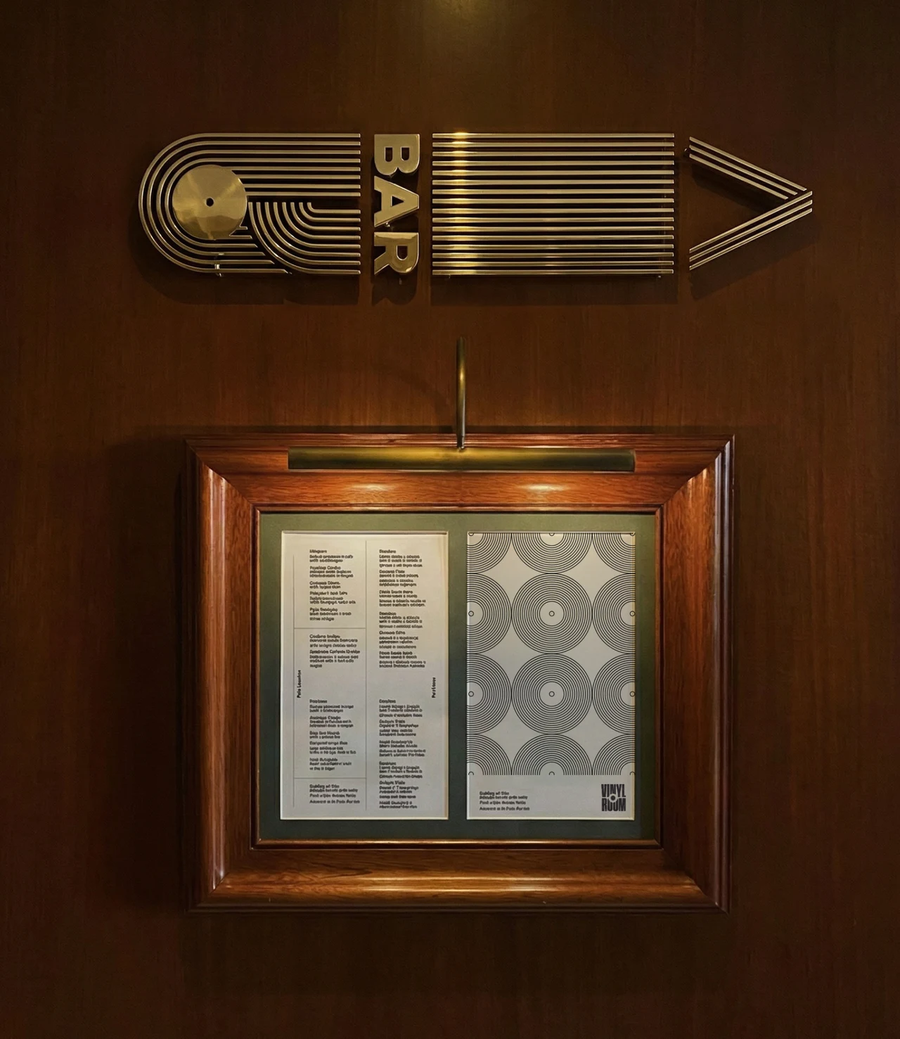 Brass Bar directional sign shaped like looping lines with a record and an arrow, above a framed menu with a record pattern lit by a brass picture light.