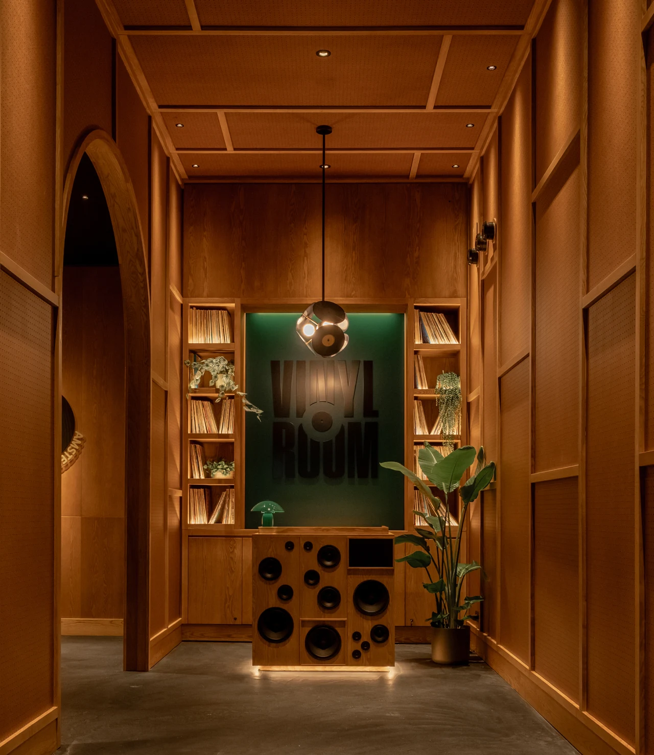 Warm wood corridor with an arch leading to a DJ booth with a speaker console, a green panel with the Vinyl Room wordmark, record shelves, plants and a record-shaped pendant.