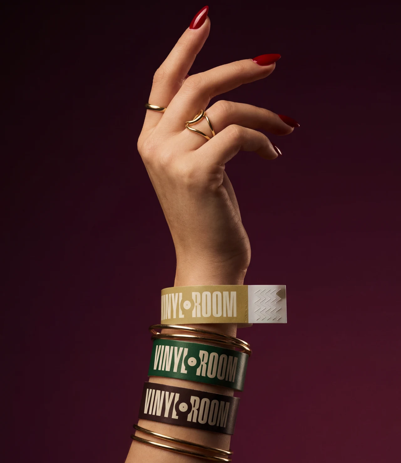 Raised hand with red nails and gold rings wearing gold, green and burgundy Vinyl Room wristbands with gold bangles, against a burgundy background.