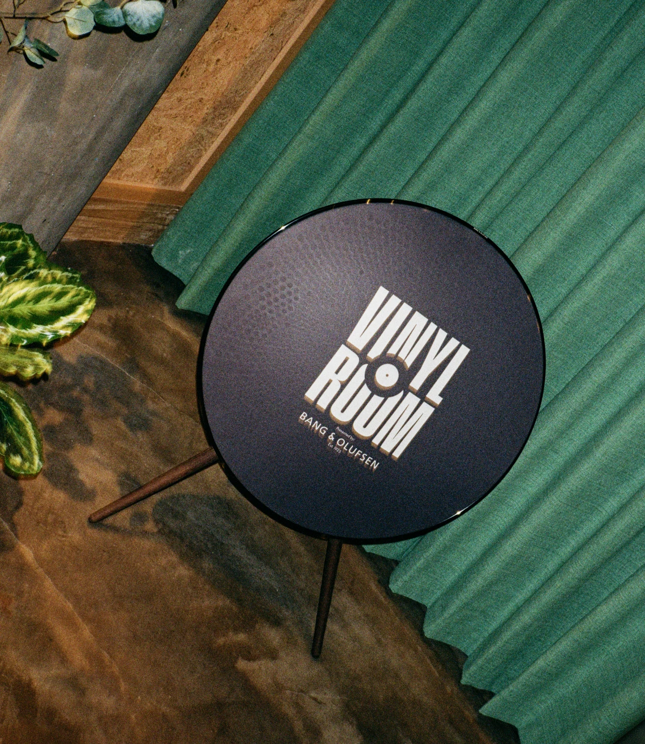 Top view of a round Bang & Olufsen speaker branded with the Vinyl Room logo, on a tripod stand beside plants and green fabric.