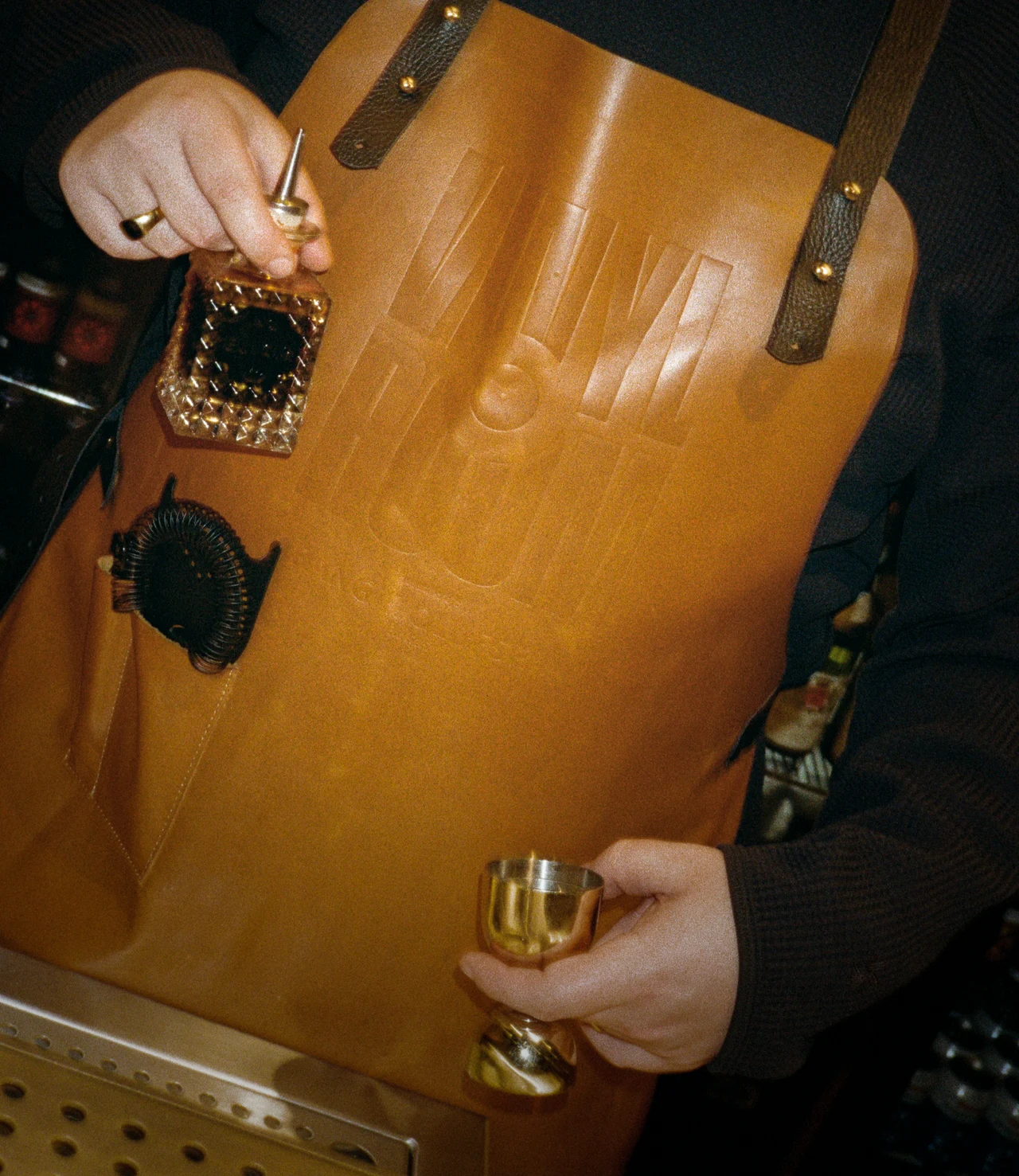 Flash photo of a bartender in a tan leather apron debossed with the Vinyl Room logo, holding a crystal bottle with a pour spout and a brass jigger.